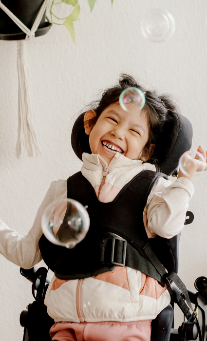 A young, smiling child with dark hair in a supportive wheelchair laughs happily as bubbles float around their face.