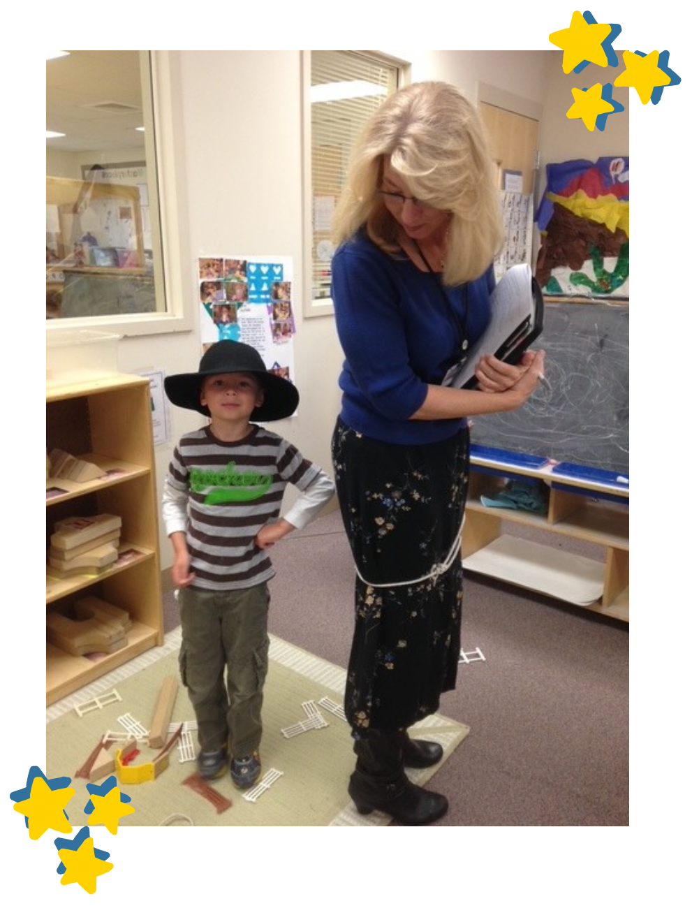 A photograph of Shelley Smith in a classroom, looking down at a young boy who is wearing a large black hat and standing with his hands on his hips. Shelley is wearing a blue sweater and a floral skirt while holding a clipboard. The photo is framed with a white border and features clusters of hand-drawn yellow stars with blue outlines in the top-right and bottom-left corners.
