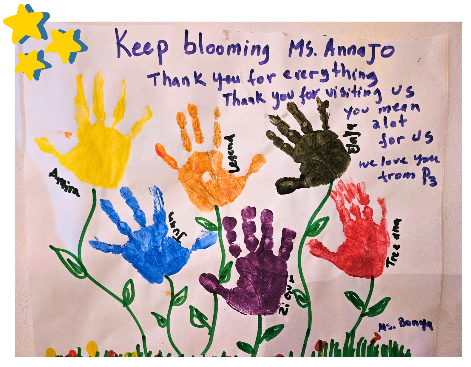A colorful handmade "Thank You" poster for Ms. Anna Jo. The poster features six handprints in yellow, orange, blue, purple, red, and black, painted to look like flowers with green stems and leaves. Each handprint is labeled with a child's name, including Amira, Legend, and Juan. Handwritten text at the top reads, "Keep blooming Ms. Anna Jo. Thank you for everything. Thank you for visiting us. You mean a lot to us. We love you from P3."
