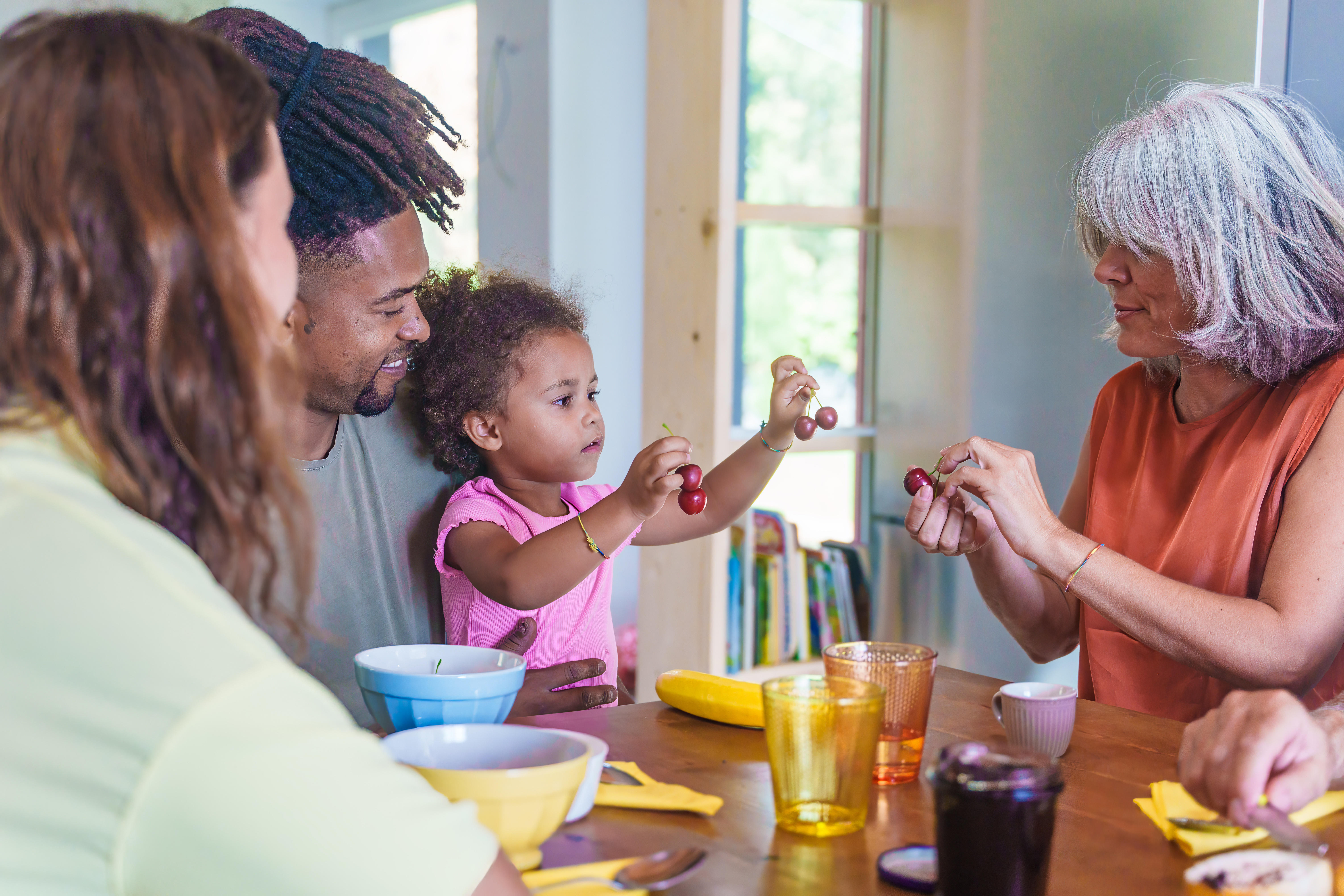 A diverse family, including a young child and an older woman, sitting around a table with bowls and drinks, interacting with cherries.